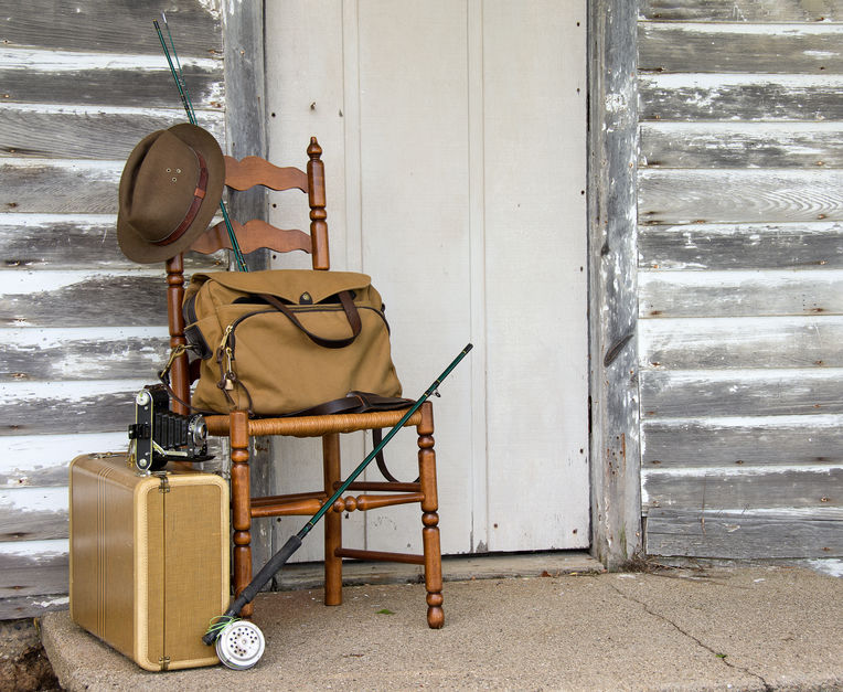 A bag, suitcase, and hat on a chair outside of a lodge. There is a fishing pole leaning on the chair.